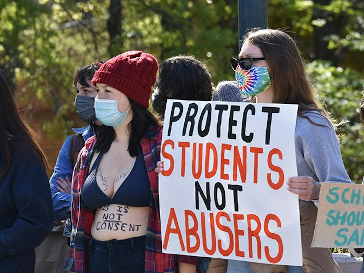 A protestor on Pickens Street Bridge holds a sign that reads, “PROTECT STUDENTS NOT ABUSERS,” and another has written, “This is not consent” and “hands off” on their body in support of the former students that accused professors of assault.