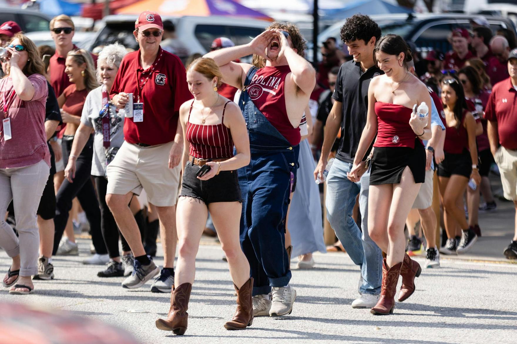 A crowd of people wearing colors like garnet, black and white in support of the University of South Carolina's football team cross the street as they walk into Williams-Brice Stadium. Most attendees in the photo are focused on one person who is cupping his hands around his mouth and yelling upward into the air. 