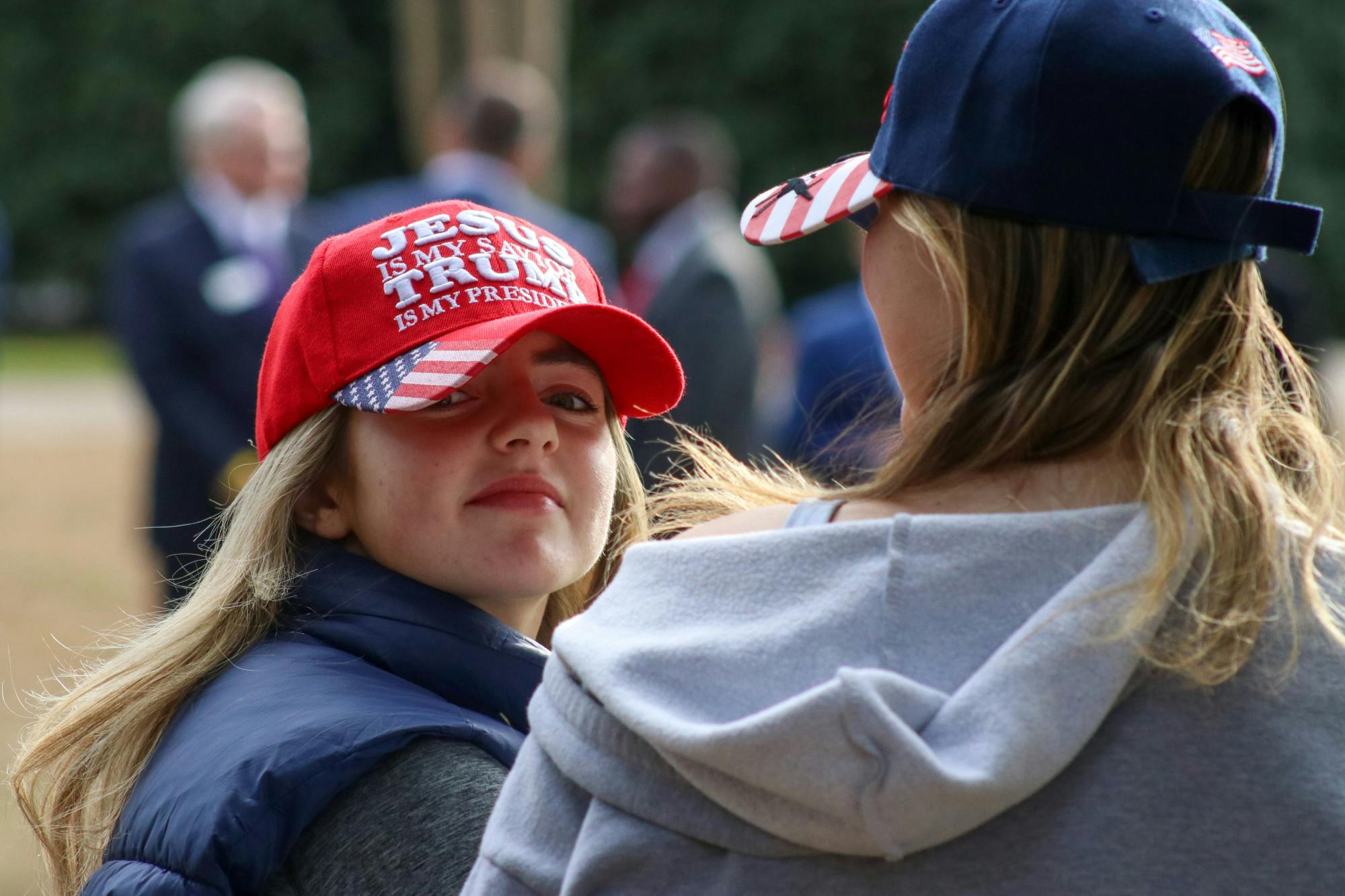 A woman (left) wears as a hat that says "JESUS is my savior, TRUMP is my president," while standing along the fence outside the Statehouse on Jan. 28, 2023. The MAGA branding of bright red and American flags could be seen across the Statehouse lawn, as former President Donald Trump's private campaign event brought many local Trump supporters back into the public eye.&nbsp;