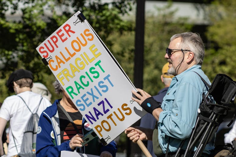 A middle-aged white man holds a sign that reads "Super Callous Fragile Racist Sexist Nazi POTUS" in ROYGBIV colors. 