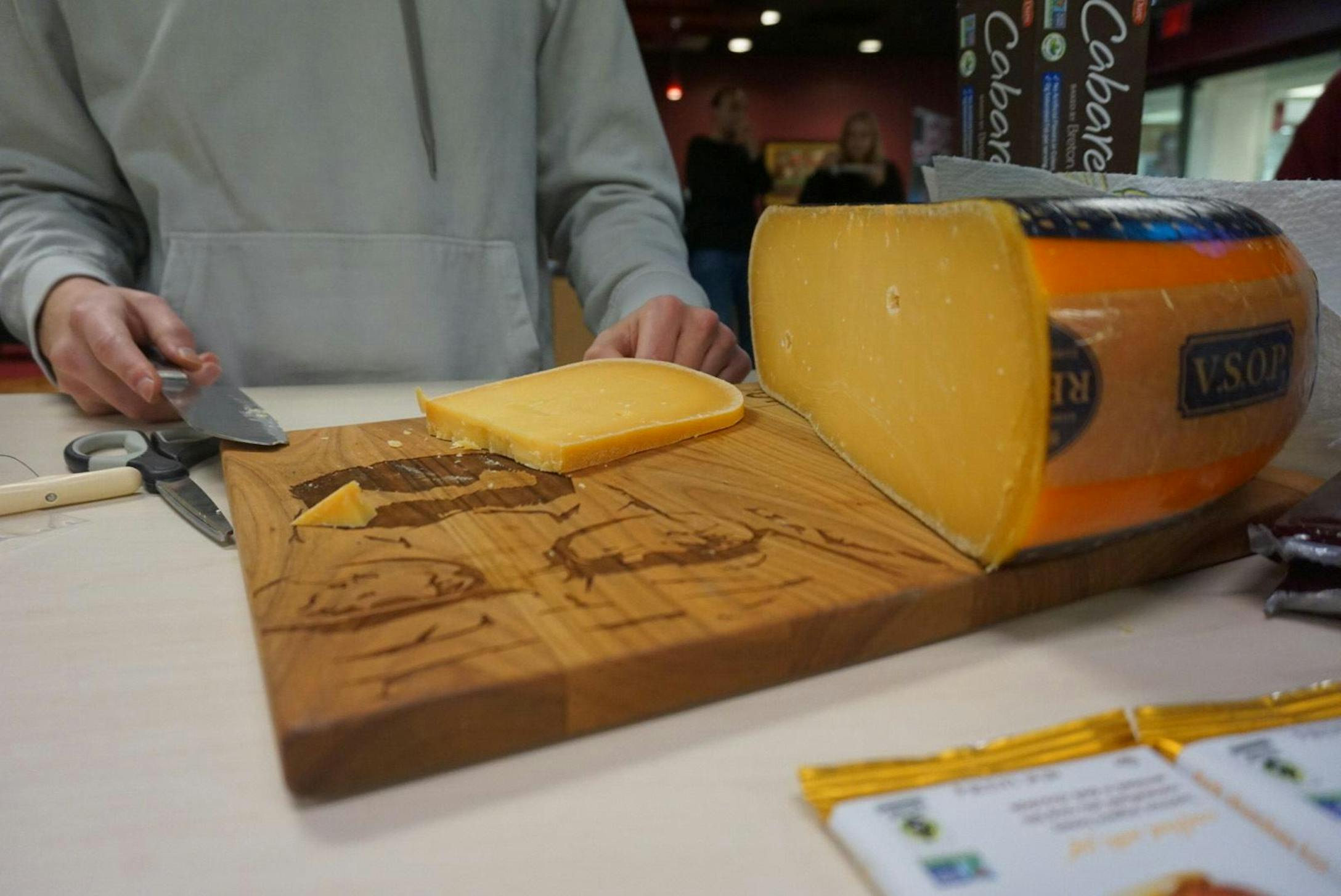 A South Carolina student wearing a light grey hooded sweatshirt stands behind a cutting board holding a large wheel of gouda cheese in orange and black wax wrapping. 