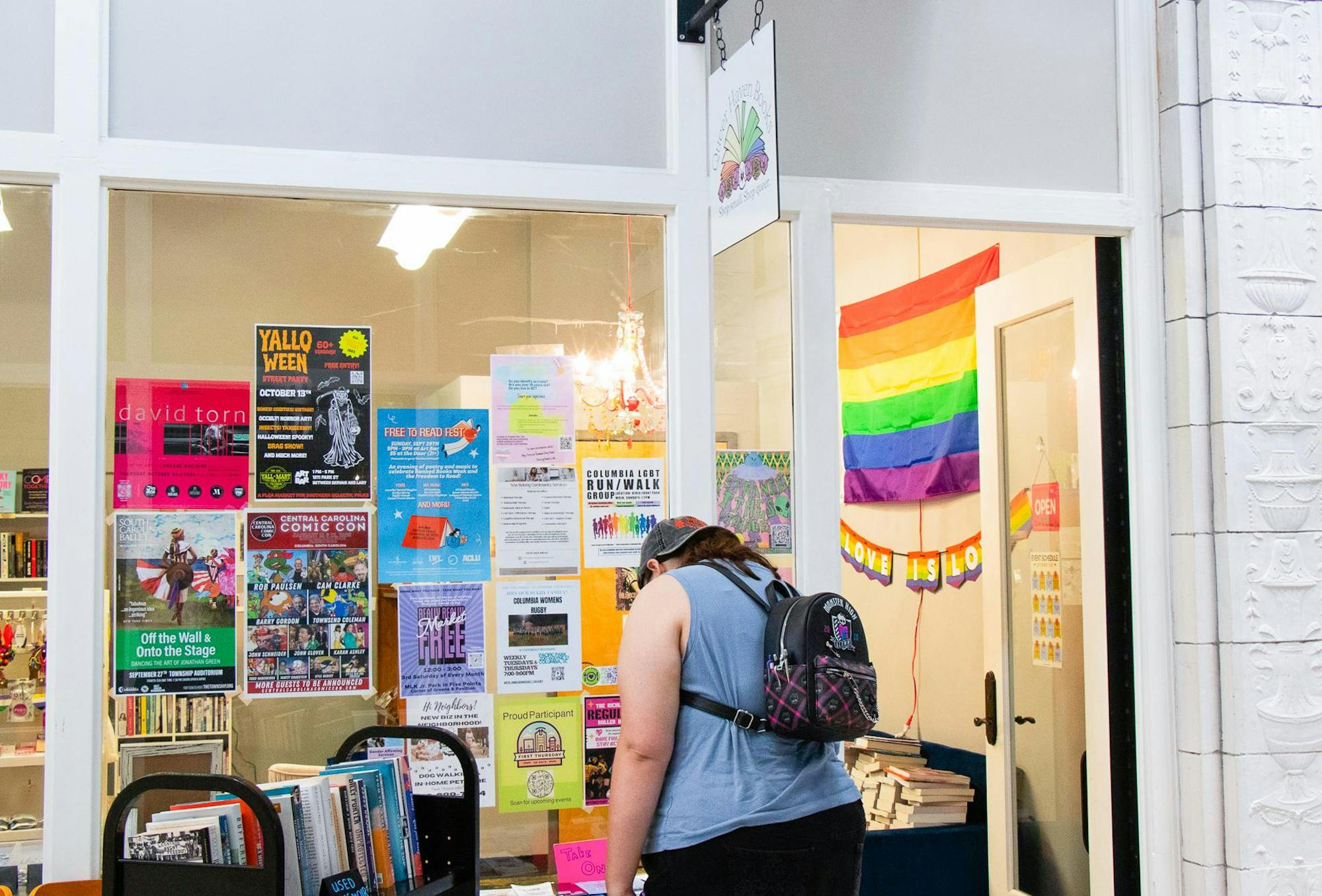 A person stands outside of the queer haven bookstore, looking at shelves of books for sale. On the window of the store is a multitude of posters advertising things such as Columbia Comic Con, Columbia LGBTQ run/walk and Yallo Ween. Inside the door of the bookstore there is a pride flag hanging on the wall. 