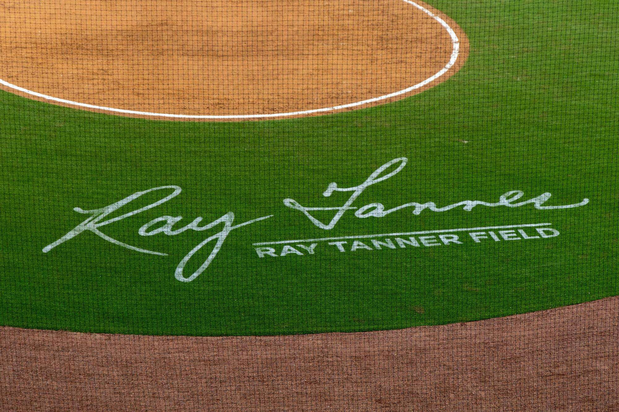 A green baseball field behind a circle of dirt, where home plate is. The field is painted in white, reading "Ray Tanner Field" with a signature.