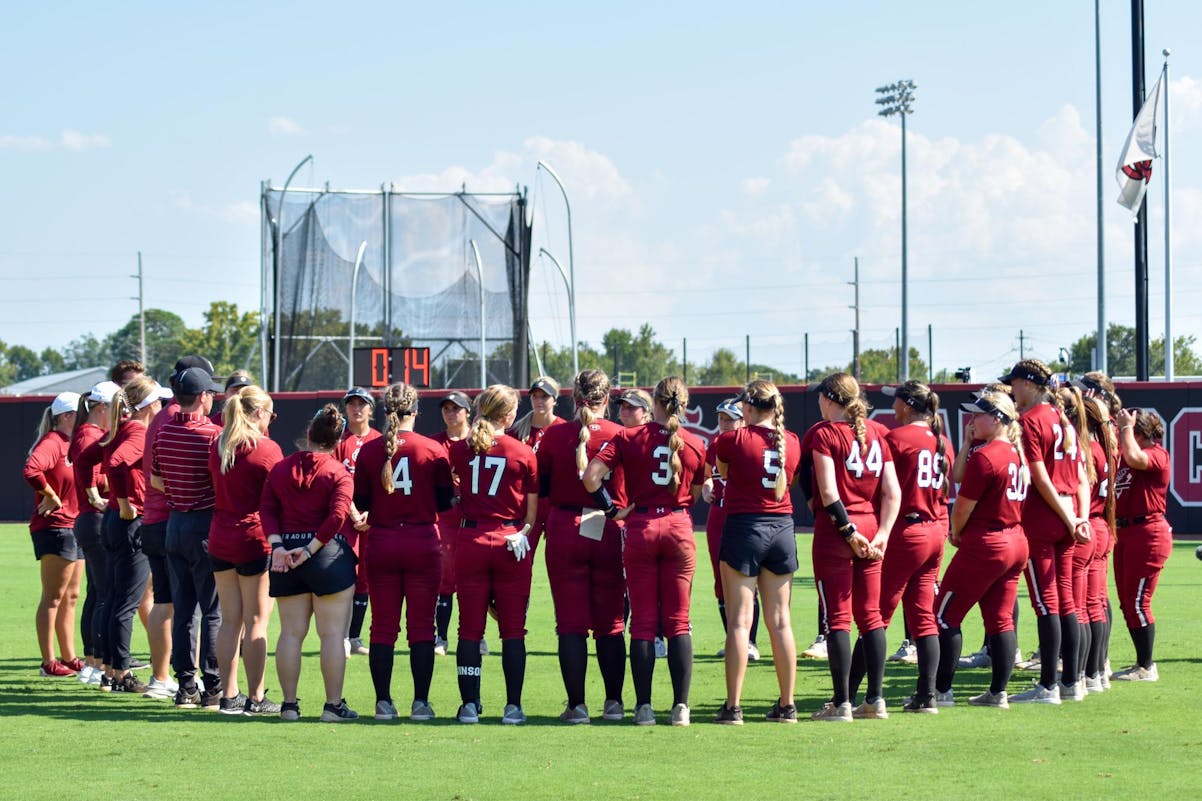 A team of South Carolina softball players, wearing red and black uniforms, stand together in a circle on the green grass outfield after playing an exhibition game. 