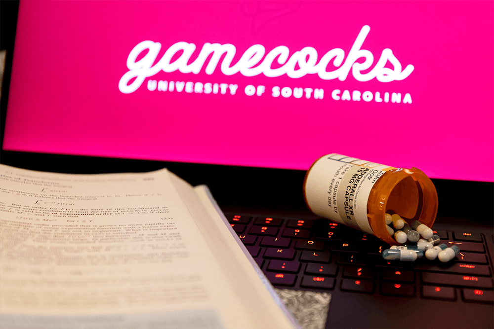 A textbook and bottle of Adderall spilled on the keyboard of a laptop. Adderall is a prescribed drug used to increase attentiveness and to help stay focused on tasks.