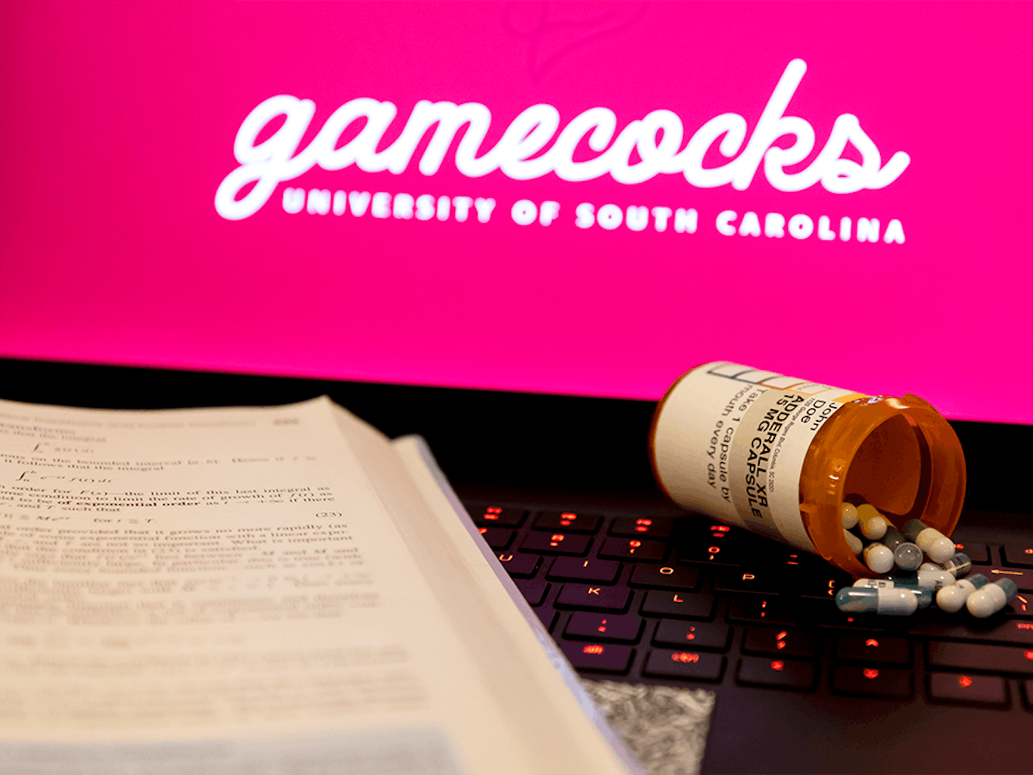 A textbook and bottle of Adderall spilled on the keyboard of a laptop. Adderall is a prescribed drug used to increase attentiveness and to help stay focused on tasks.