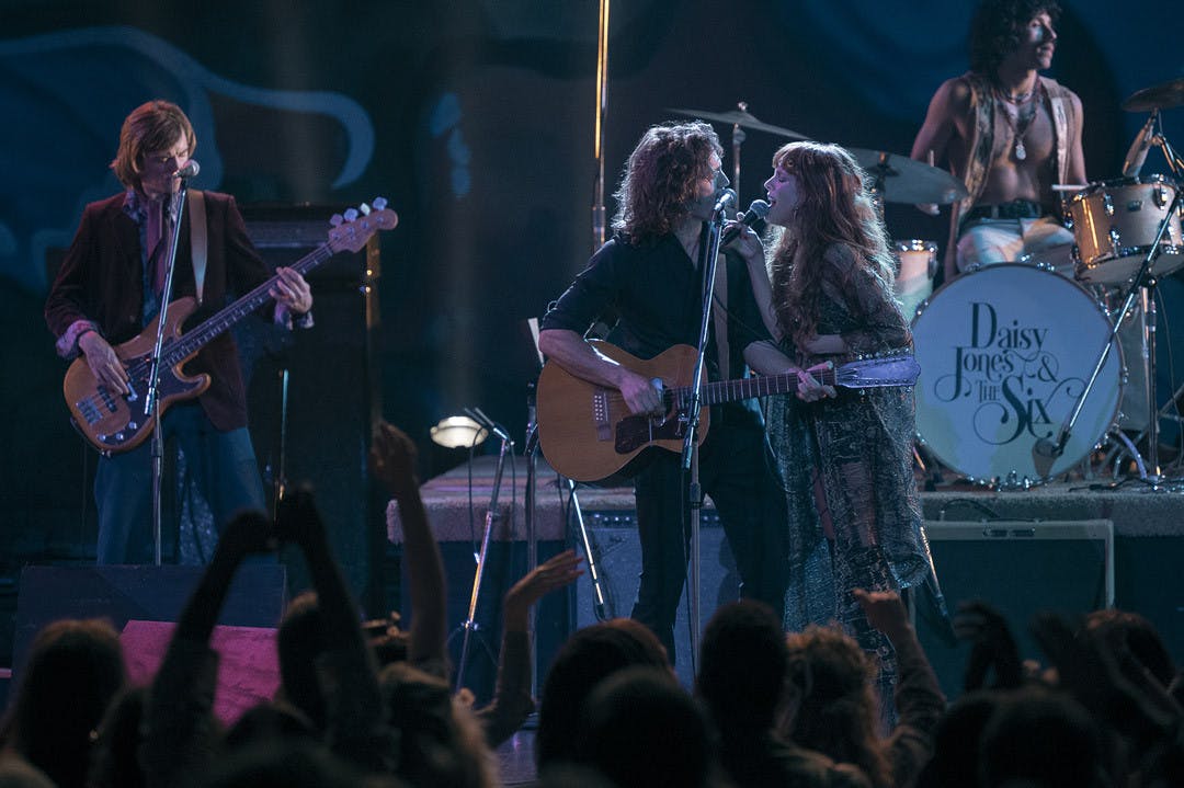 Sam Claflin (center) and Riley Keough (right) sing together during a scene from the drama miniseries, &nbsp;"Daisy Jones &amp; The Six." The series is a live-action adaptation of American author Taylor Jenkins Reid's novel by the same name, and it follows the lives of a fictional 1970s rock band from the perspective of their older selves.&nbsp;