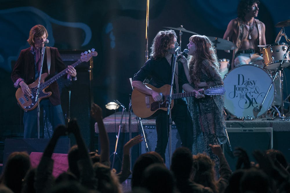 <p>Sam Claflin (center) and Riley Keough (right) sing together during a scene from the drama miniseries, &nbsp;"Daisy Jones &amp; The Six." The series is a live-action adaptation of American author Taylor Jenkins Reid's novel by the same name, and it follows the lives of a fictional 1970s rock band from the perspective of their older selves.&nbsp;</p>