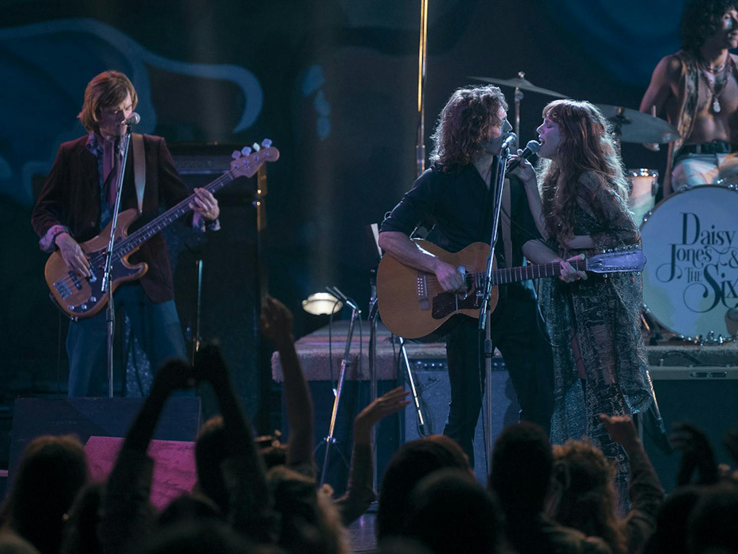 Sam Claflin (center) and Riley Keough (right) sing together during a scene from the drama miniseries, &nbsp;"Daisy Jones &amp; The Six." The series is a live-action adaptation of American author Taylor Jenkins Reid's novel by the same name, and it follows the lives of a fictional 1970s rock band from the perspective of their older selves.&nbsp;