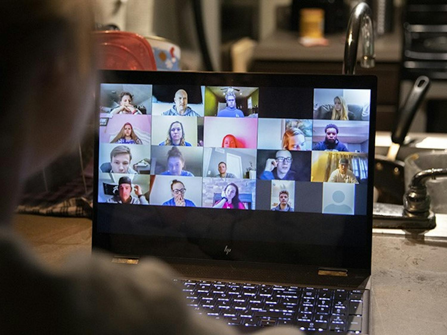 A person sits in their kitchen while watching a pre-recorded Zoom lecture.