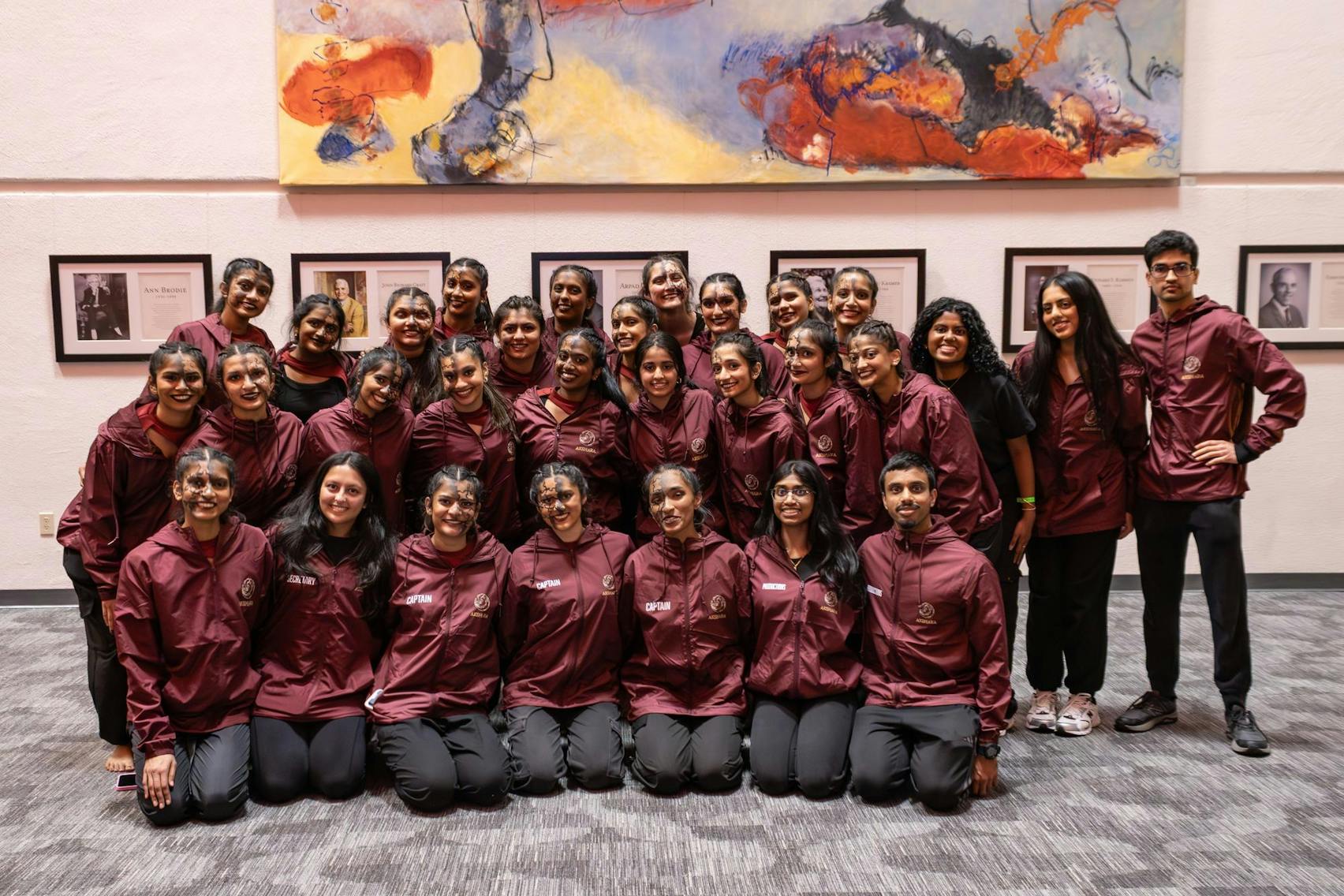 Members of a dance team wearing matching maroon jackets pose together in several rows inside a building, smiling after a performance, with framed portraits and a large abstract painting on the wall behind them.