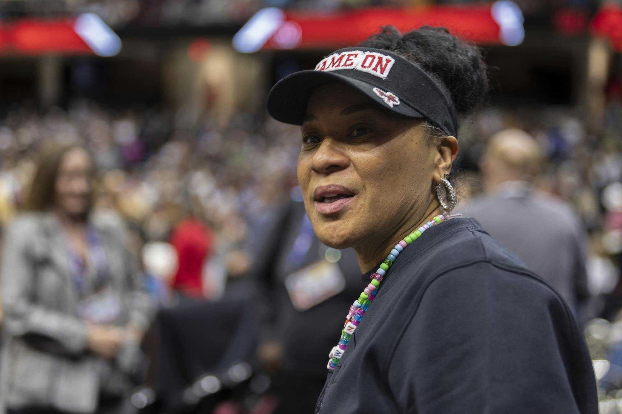 A close-up shot of Head Women's Basketball Coach Dawn Staley in front of a crowd of people. Coach Staley is wearing a black sweatshirt, a black Gamecocks visor that reads "game on" and a beaded necklace. 