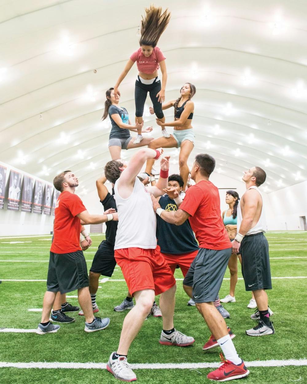 The Large Co-Ed Cheerleading Team practices rewinds at the Football Indoor Practice Facility during an early morning practice. Two of the men, bottom-center, throw the top girl in a backward spin to the middle women, center, for the catch while the bases brace under the weight and spotters ensure the safety of all involved.
