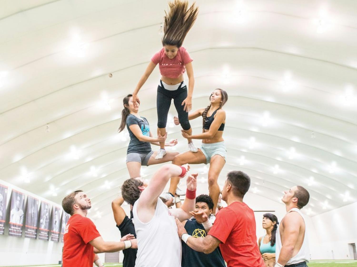 The Large Co-Ed Cheerleading Team practices rewinds at the Football Indoor Practice Facility during an early morning practice. Two of the men, bottom-center, throw the top girl in a backward spin to the middle women, center, for the catch while the bases brace under the weight and spotters ensure the safety of all involved.