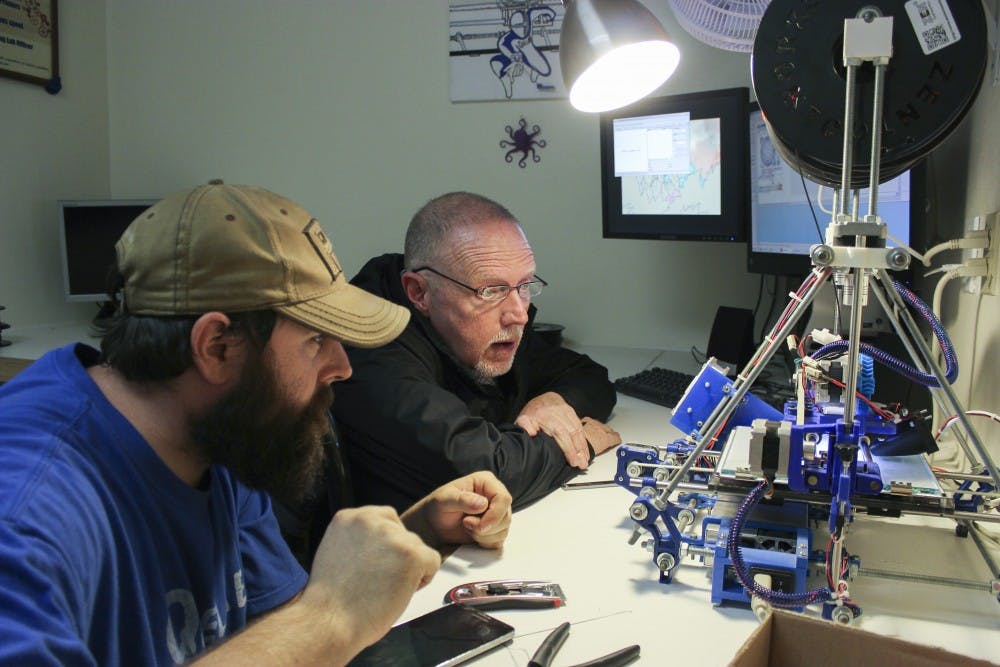 John Cochrane, left, and Ben Cole wait for a design to form on the 3-D printer at QueLab on March 10. QueLab aims to bring locals together to innovate ideas on technology and to use the facilities to work on personal projects.