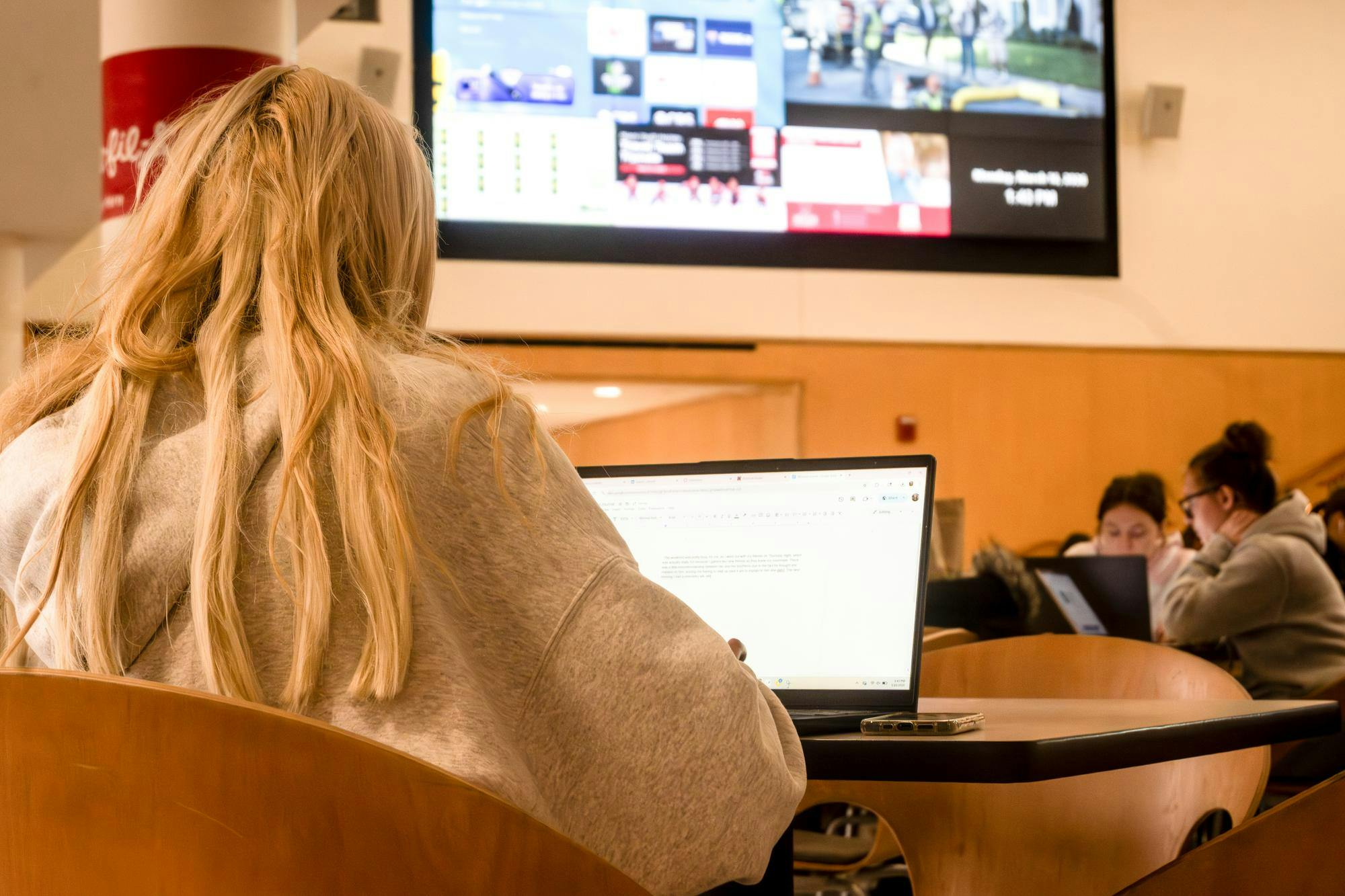 A student types on their computer in Armstrong Student Center while a TV plays in the background.