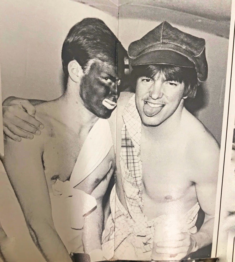 On the fold between two pages of the 1982 edition of Recensio, two white members of Alpha Epsilon Pi link arms at a party. The face in the middle of the fold is covered in black paint, with bright white around his mouth.