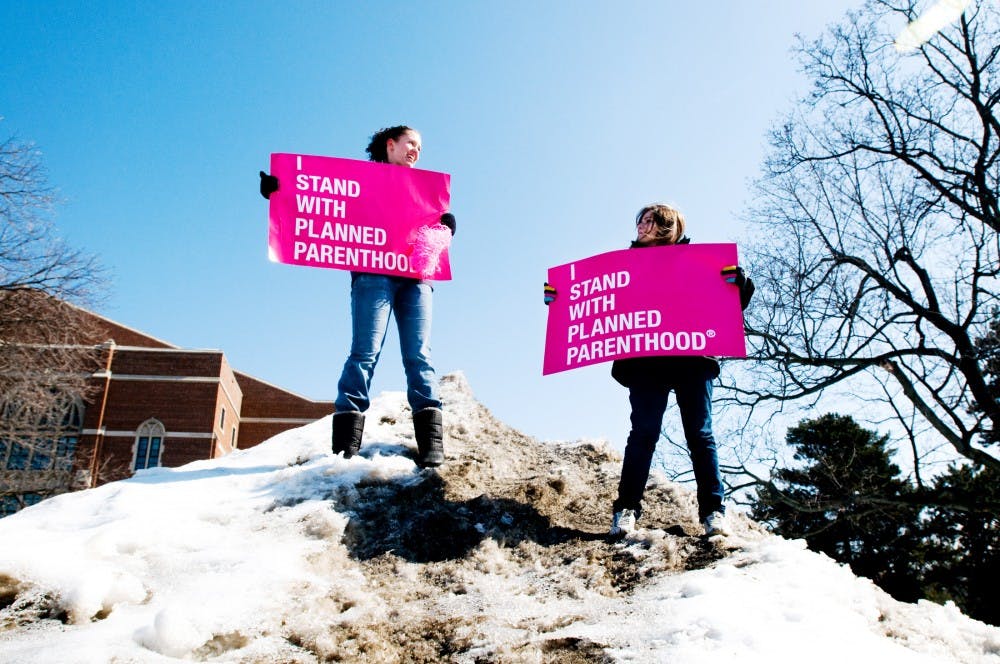Biosystems engineering junior Maureen Berryman, left, and general management junior Samantha Bowles hold signs supporting Planned Parenthood while standing on an ice mound near the rock on Farm Lane Tuesday afternoon during a protest of the funding cuts proposed for Planned Parenthood. The Students for a Choice group hosted the protest, and had people sign a petition supporting the public organization, which provides services like cancer screening and birth control to women. Lauren Wood/The State News