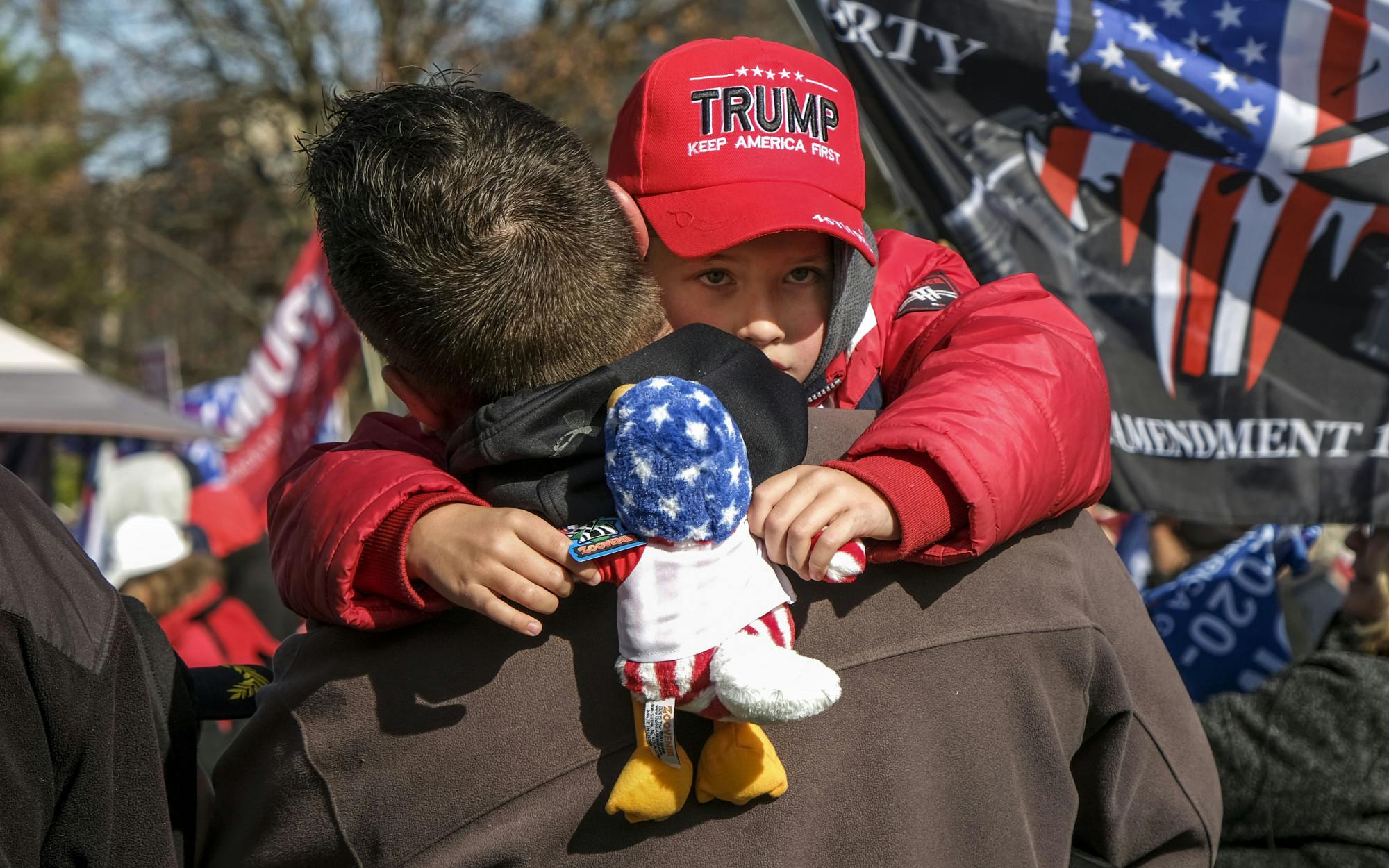 A young child wearing a Trump hat holds a stuffed animal while being carried by another person.