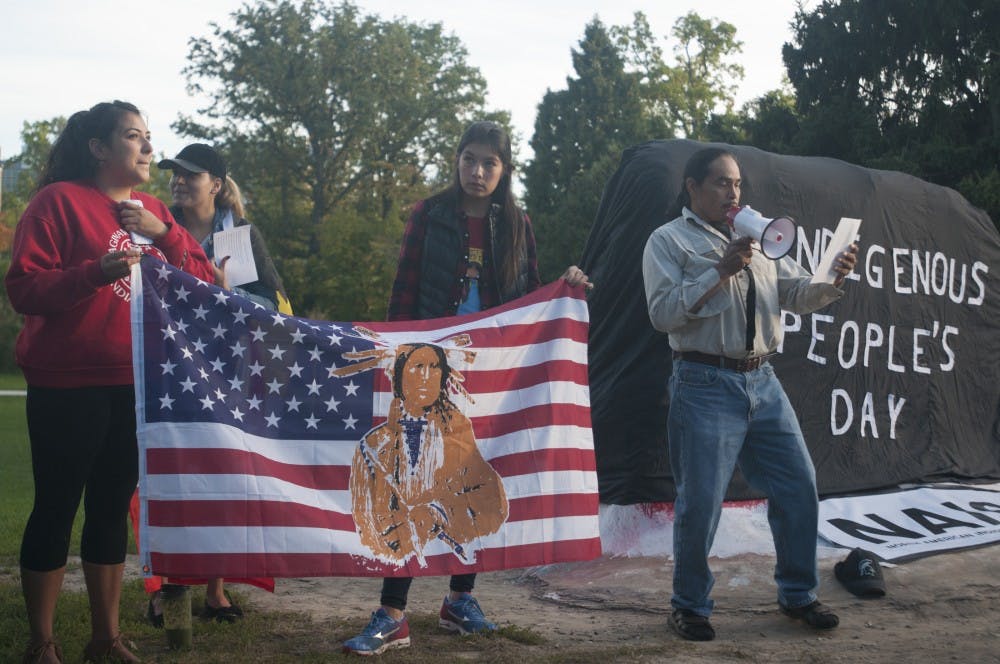 From left, world politics and humanities senior Cassandra Shavrnock and nutritional science senior Shelbie Shelder hold an American flag with a native man on it while Haslett resident Aarin Dokum reads an introduction to Indigenous People's Day on Oct. 10, 2016 at The Rock. Dokum read the introduction in Anishinaabemowin, the language of the Ojibwe people, and in English. 