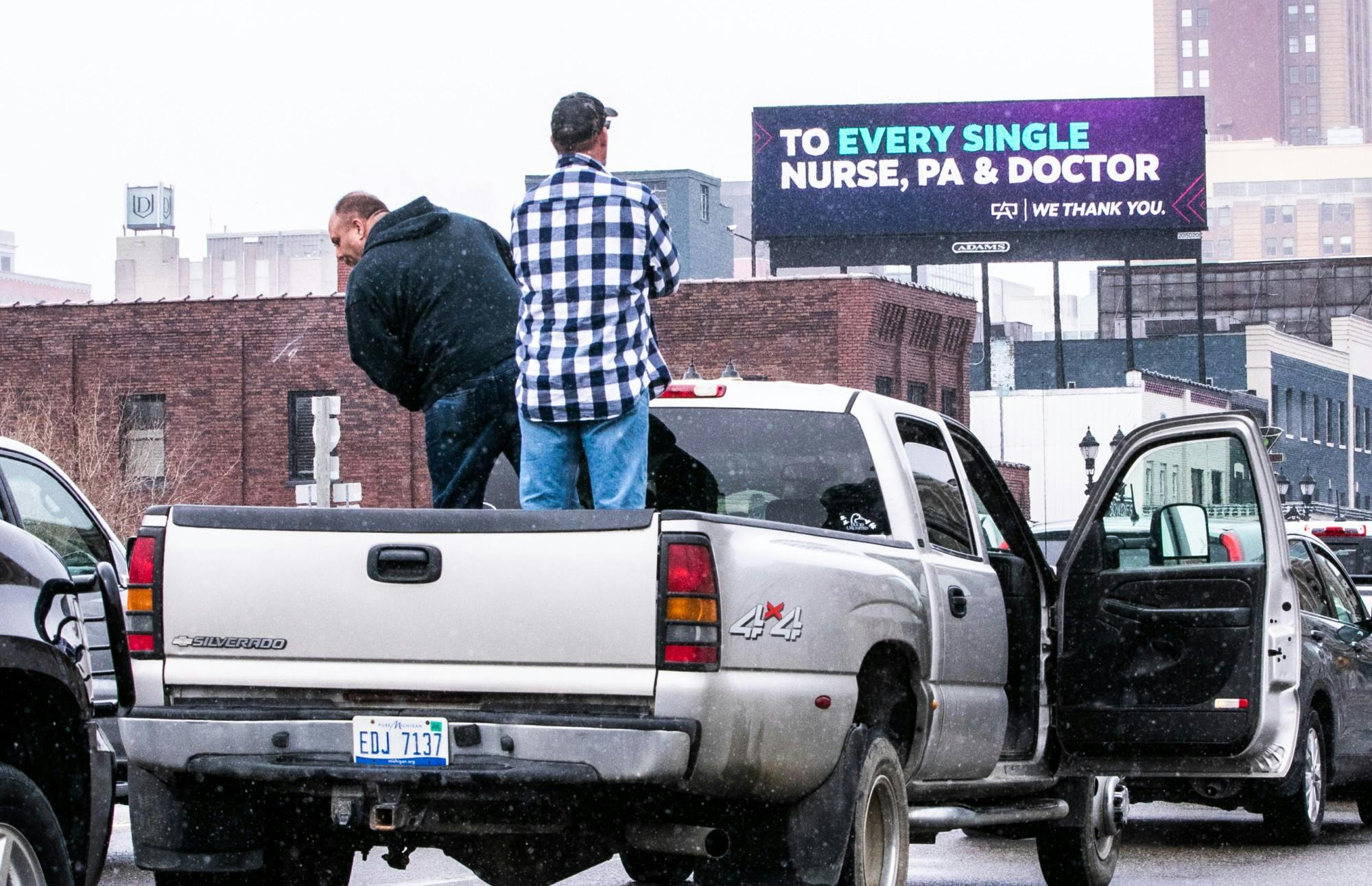 Two people stand in the back of a pickup truck. One person spits off the side of the truck. In the background of the photo is a billboard that reads, 'To every single nurse, PA & doctor, we thank you'