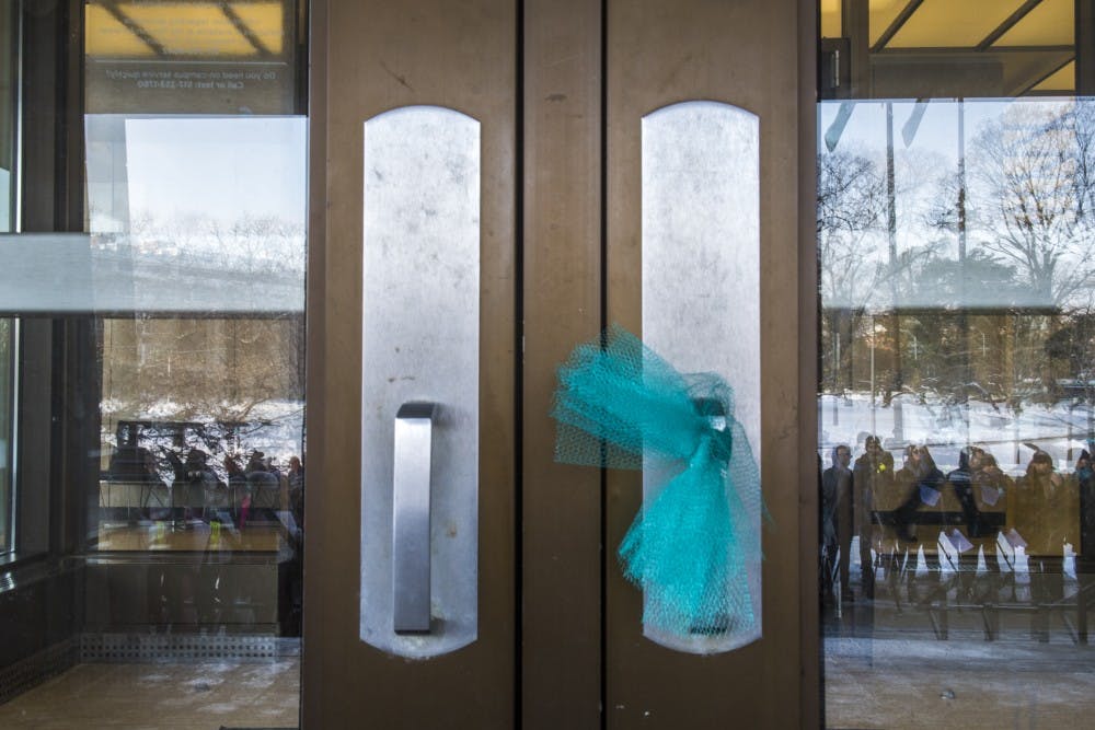 Protesters are seen through the reflection the Hannah Administration Building's doors during the MARCH on Hannah on Feb. 6, 2018. Protesters marched from Erickson Hall to the Hannah Administration Building and had a list of demands, including the resignation of the Board of Trustees and Interim President Engler. (Nic Antaya | The State News)