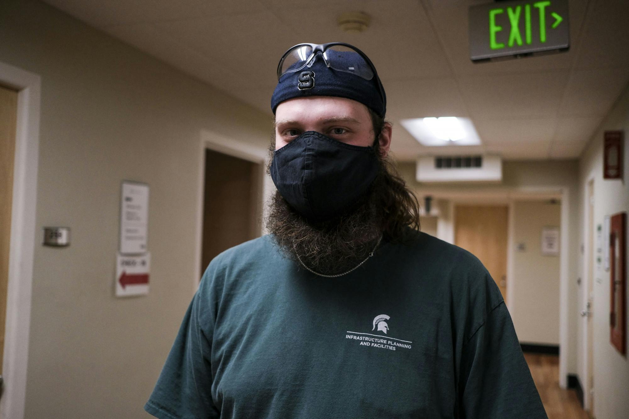 A bearded person wearing a face mask and a backwards cap with eye protection glasses resting on it stands in a hallway posing for a picture.