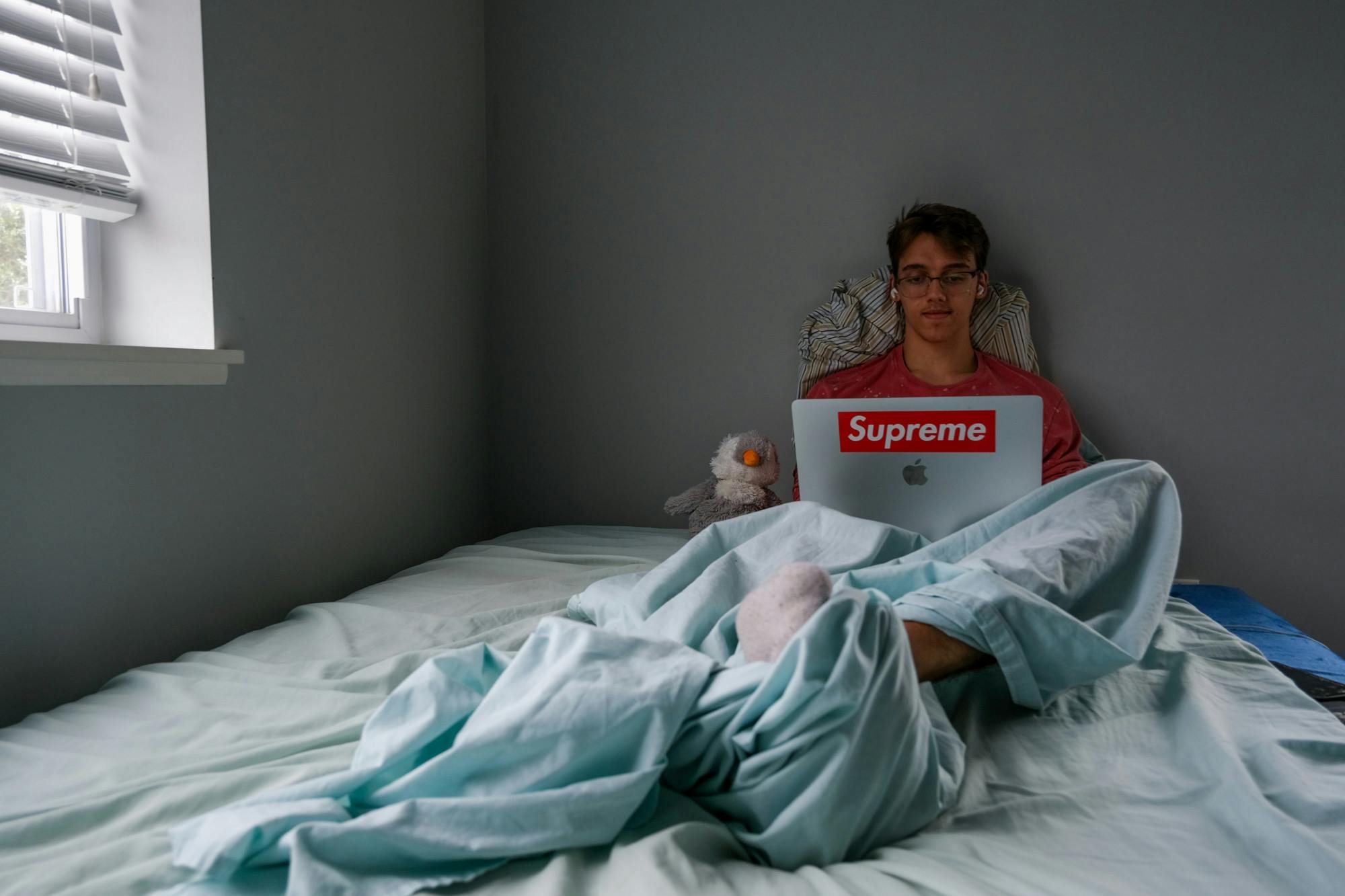 A person sits on a bed and works on a laptop with a Supreme sticker on it.