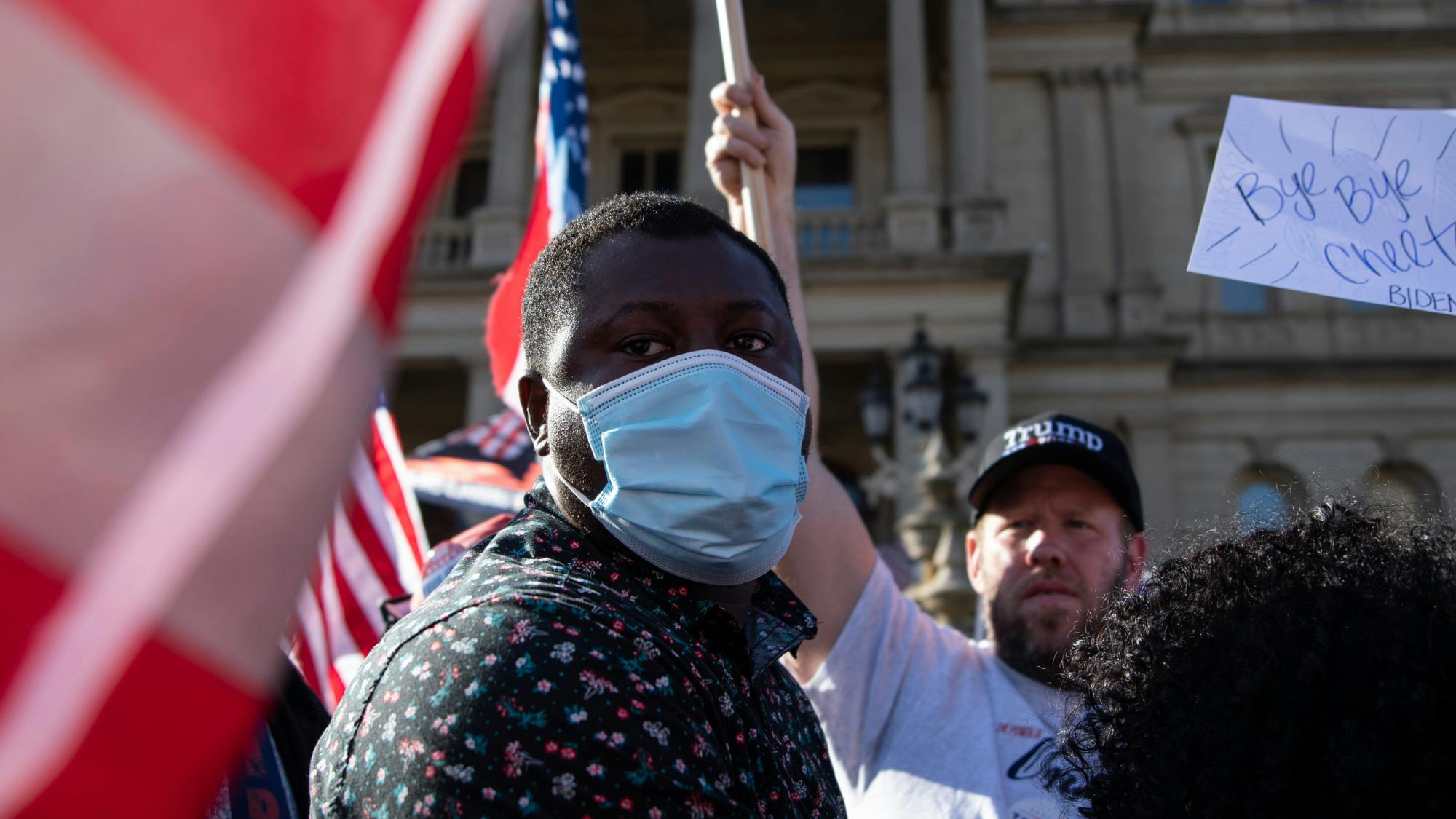 A person wearing a face mask stands in the foreground while a person wearing a Trump hat stands in the background.