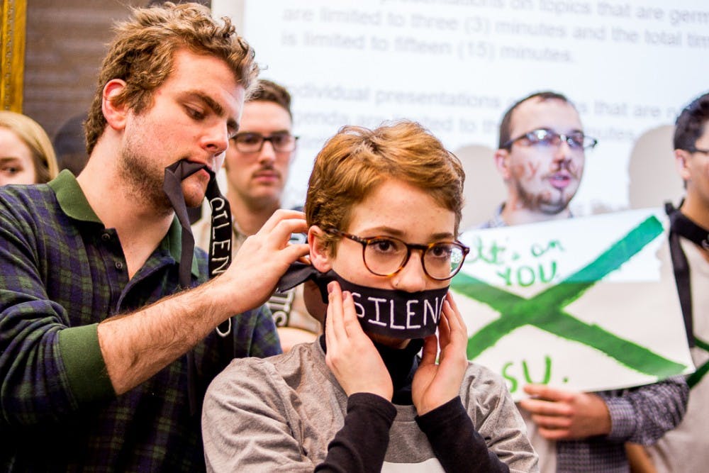 Drew Cook helps Morgan McCaul, a victim of Nassar’s abuse tie a gag that reads "silence" around her mouth  during a Board of Trustees meeting on Dec 15, 2017 at the Hannah Administration Building. Protesters gathered to address the misconduct of MSU’s handling of the Larry Nassar investigation. 