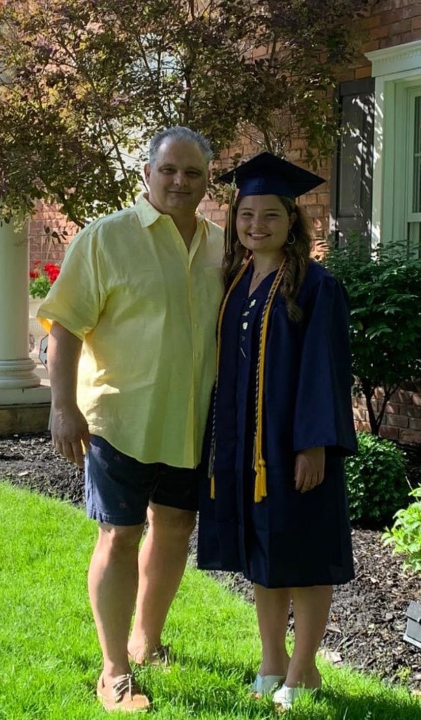 A middle-aged man and a young girl stand and pose for a photo in a yard. The young girl wears a navy blue graduation cap and gown.