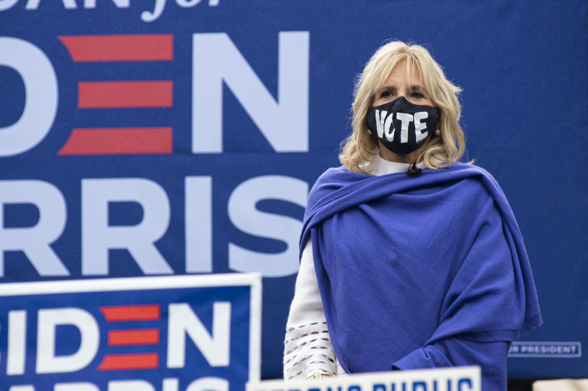 Now-First Lady Dr. Jill Biden a face mask with the word 'vote' and a shawl stands in front of a large Biden Harris sign.
