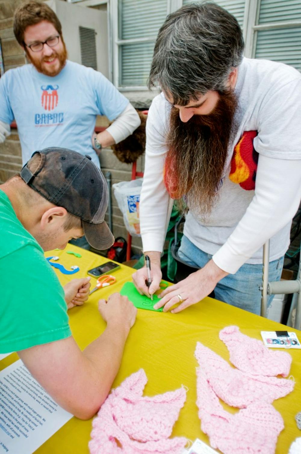 MSU alumni Ian Walker, left, and John Buckler, right, run an arts and crafts table on Saturday in downtown East Lansing as local non-profit organizations gather to raise awareness. The pair arrived representing the Great American Fierce Beard Organization as they cut paper mustaches and sold cloth beards to attendees such as Lansing resident Justin Caine, pictured. Mo Hnatiuk/The State News