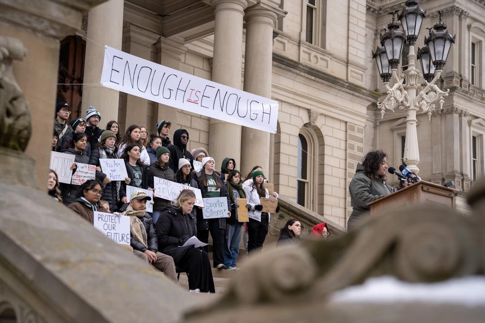 Psychology senior and activist Maya Manuel speaks at the podium while MSU students rally behind her on the steps of the Michigan State Capitol for the sit down protest against gun violence on Feb. 15, 2024. 