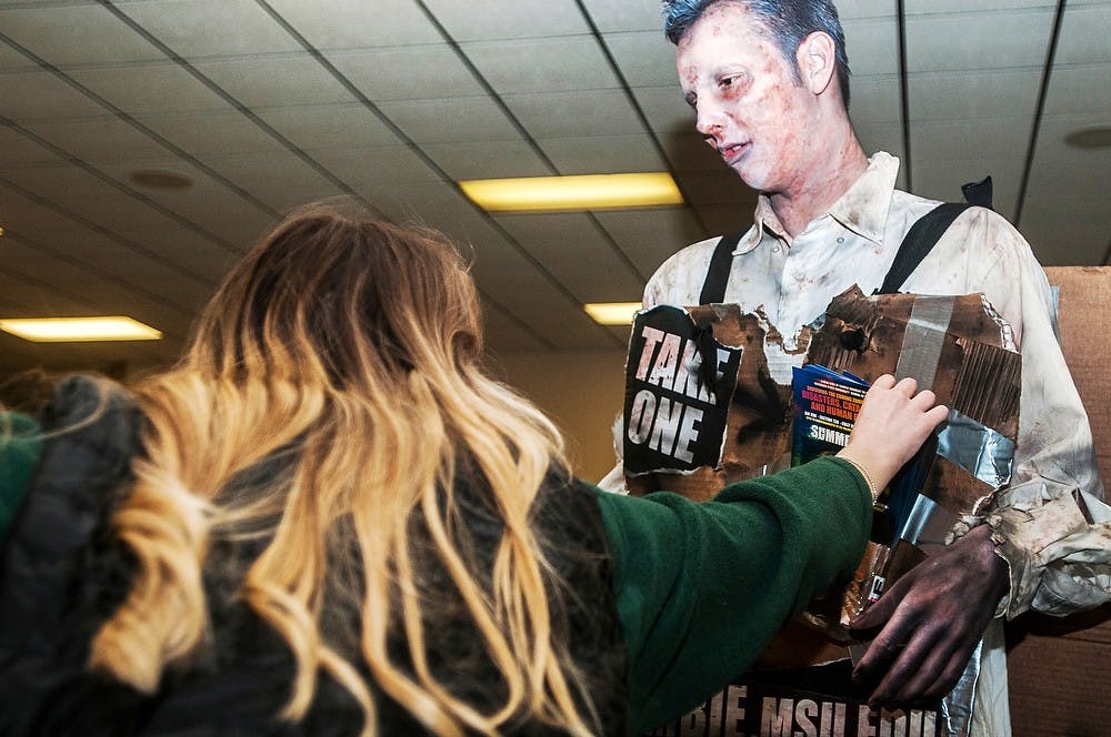 <p>Communication freshman Lauren Bain reaches to grab a flyer from criminal justice freshman Kevin Hodge who dressed as a zombie to promote the online course 