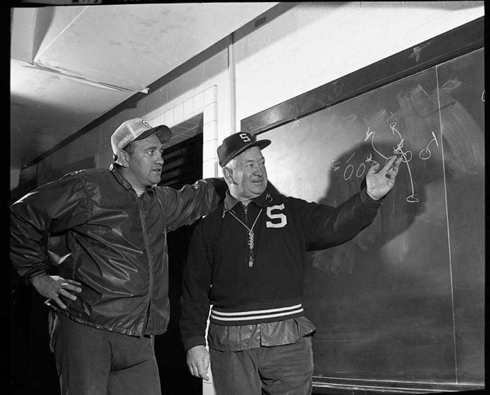 <p>Defensive coordinator Hank Bullough looks at a play diagrammed on the chalkboard with head coach Duffy Daugherty. (MSU Athletic Communications)</p>