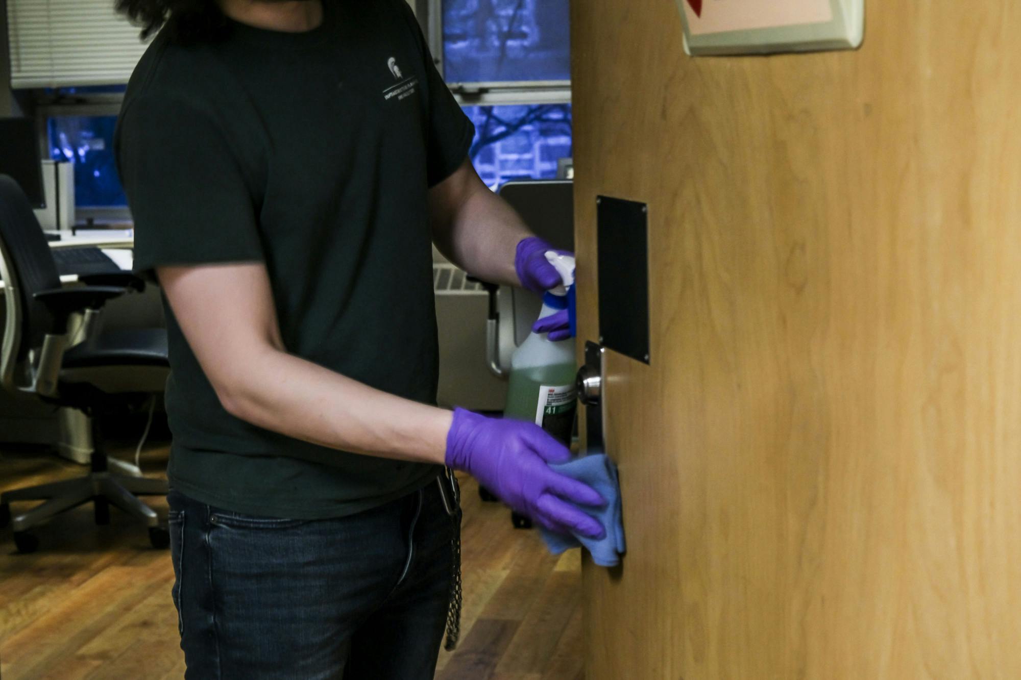 A person wearing purple medical gloves uses a rag to wipe down a doorhandle while holding a cleaning spray. The person's face is not visible.