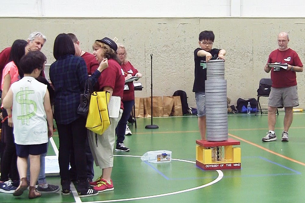 <p>A student from Chung-Ang University Elementary School, Seocho-gu, South Korea, places a weight atop a stack of them to test the durability of the balsa wood structure beneath. The student was competing in an Odyssey of the Mind event called, "Lose Your Marbles," on May 22 at IM Sports-West. Still from video: Jessica Steeley/The State News</p>