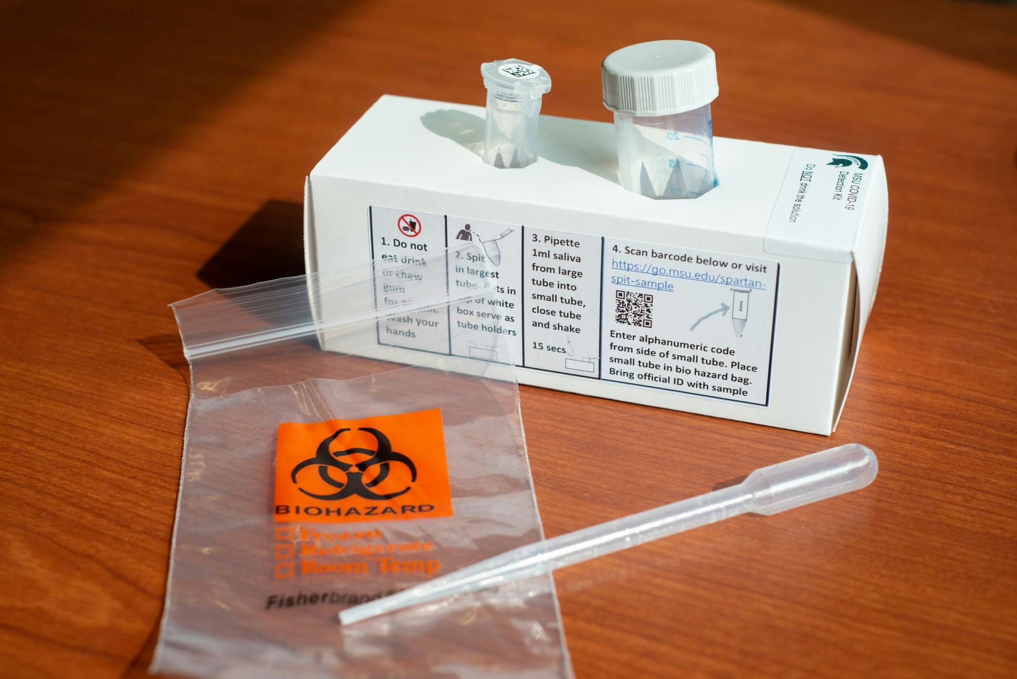 A cardboard box holds two collection tubes. In front of the box is a bag labeled 'Biohazard.'
