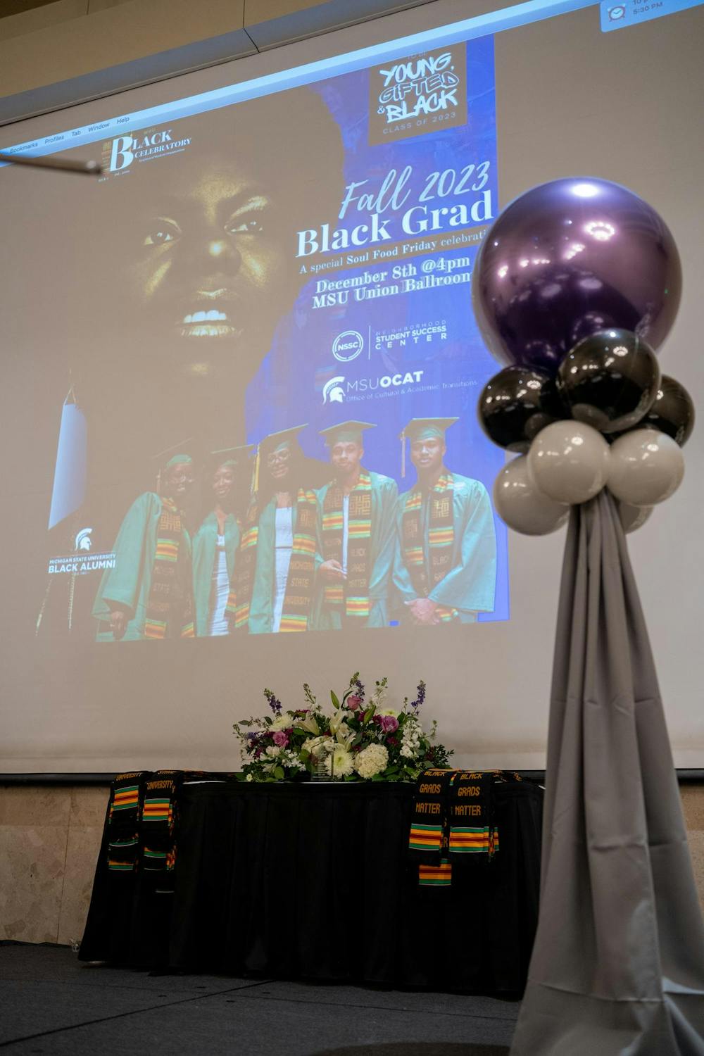 The Fall 2023 Black Grad event flyer on the screen and the “Black Grads Matter” stoles lay on a table on stage before the event at the Union Ballroom on Dec. 8, 2023. 