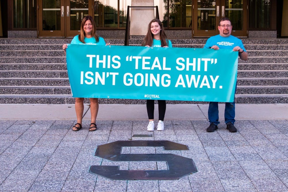 <p>Leslie Miller, Emma Ann Miller, and Bryan Tarrant (left to right) pose with a sign they brought outside of the Hannah Administration Building before &nbsp;the Board of Trustees meeting on Aug. 31, 2018. Emma Ann (daughter of Leslie) is a Nassar survivor herself, and Tarrant's daughter, Jessica Tarrant, is also a Nassar survivor.</p>