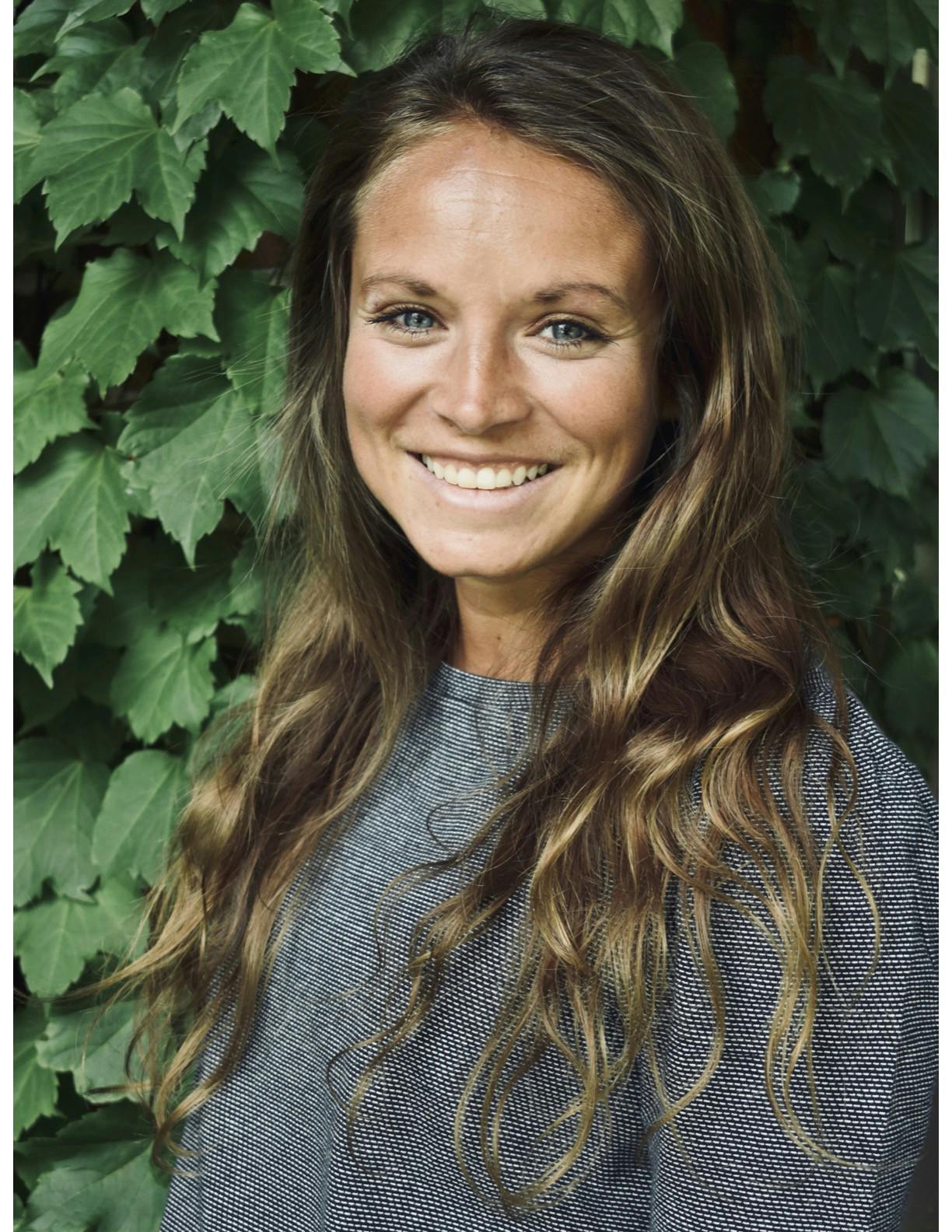 A person smiles and poses for a photo in front of a wall covered in leaves.