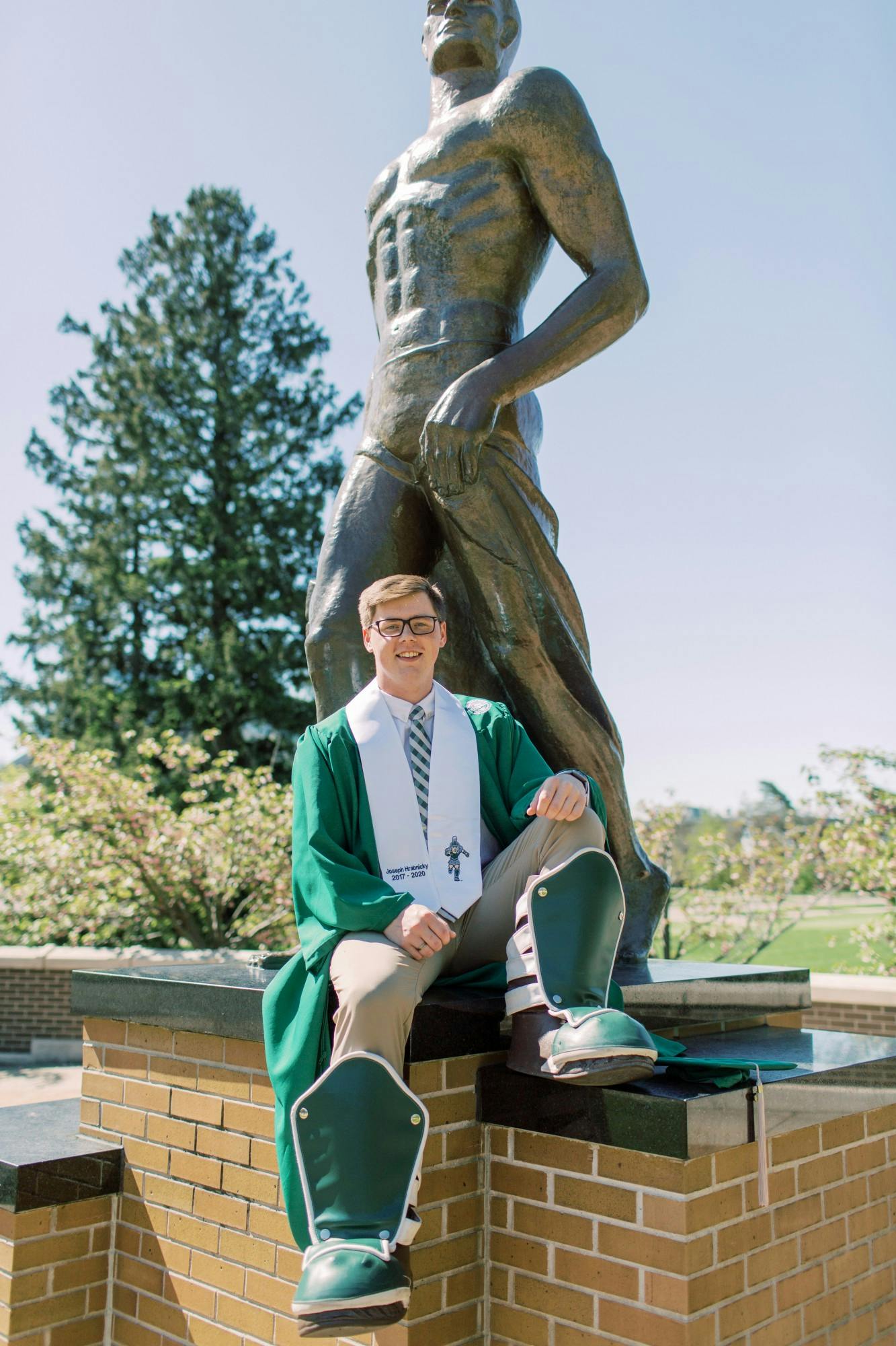 MSU alumni Joseph Hrabnicky poses in front of the Spartan Statue in his graduation gown and Sparty boots. Hrabnicky played Sparty since the second semester of his freshman year at MSU.