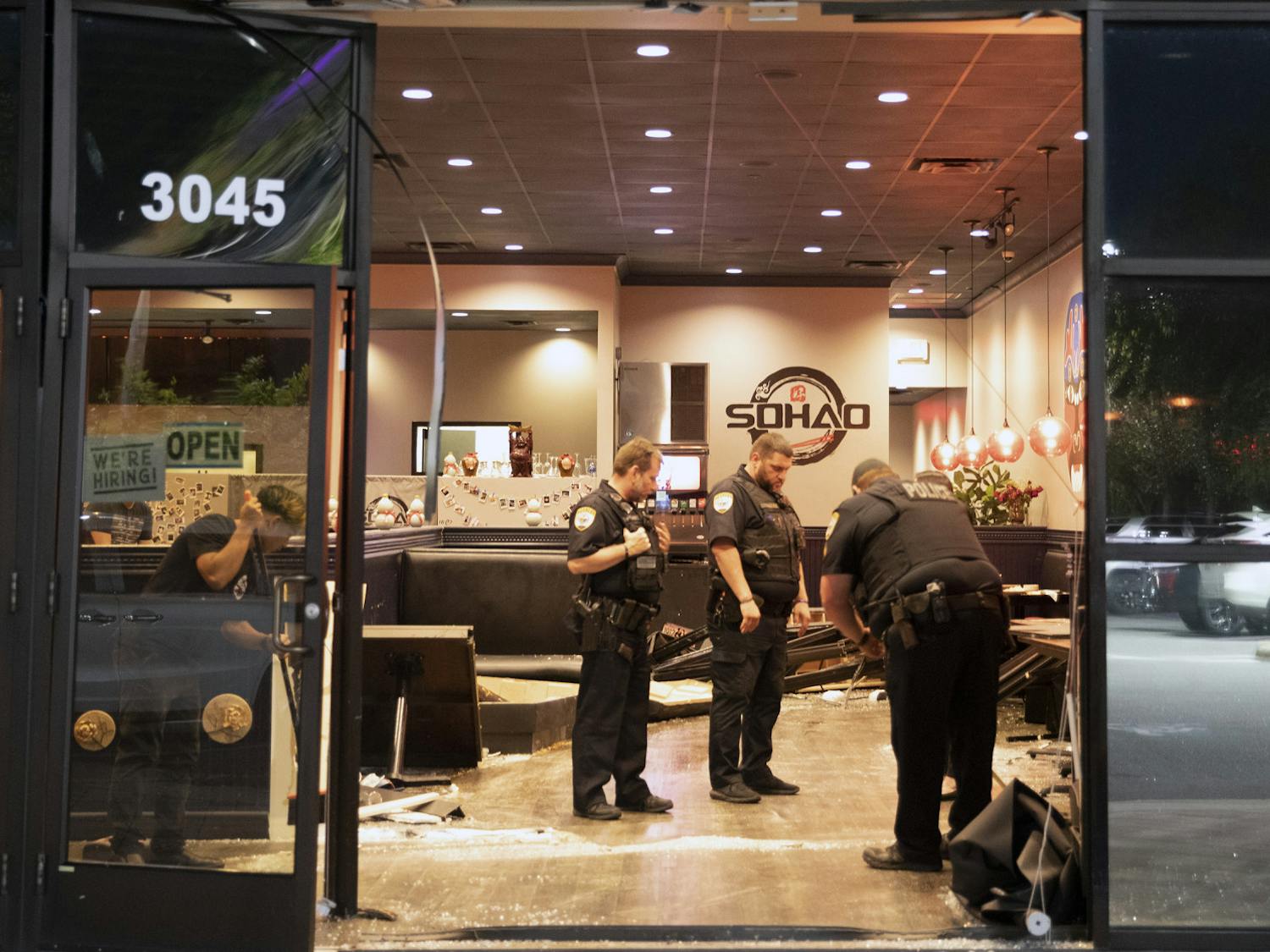 Police officers investigate the area and help clean broken glass from the floor of SOHAO Cafe & Gator Suyaki after a blue Dodge was driven through the front of the restaurant on Monday, May 17, 2021.