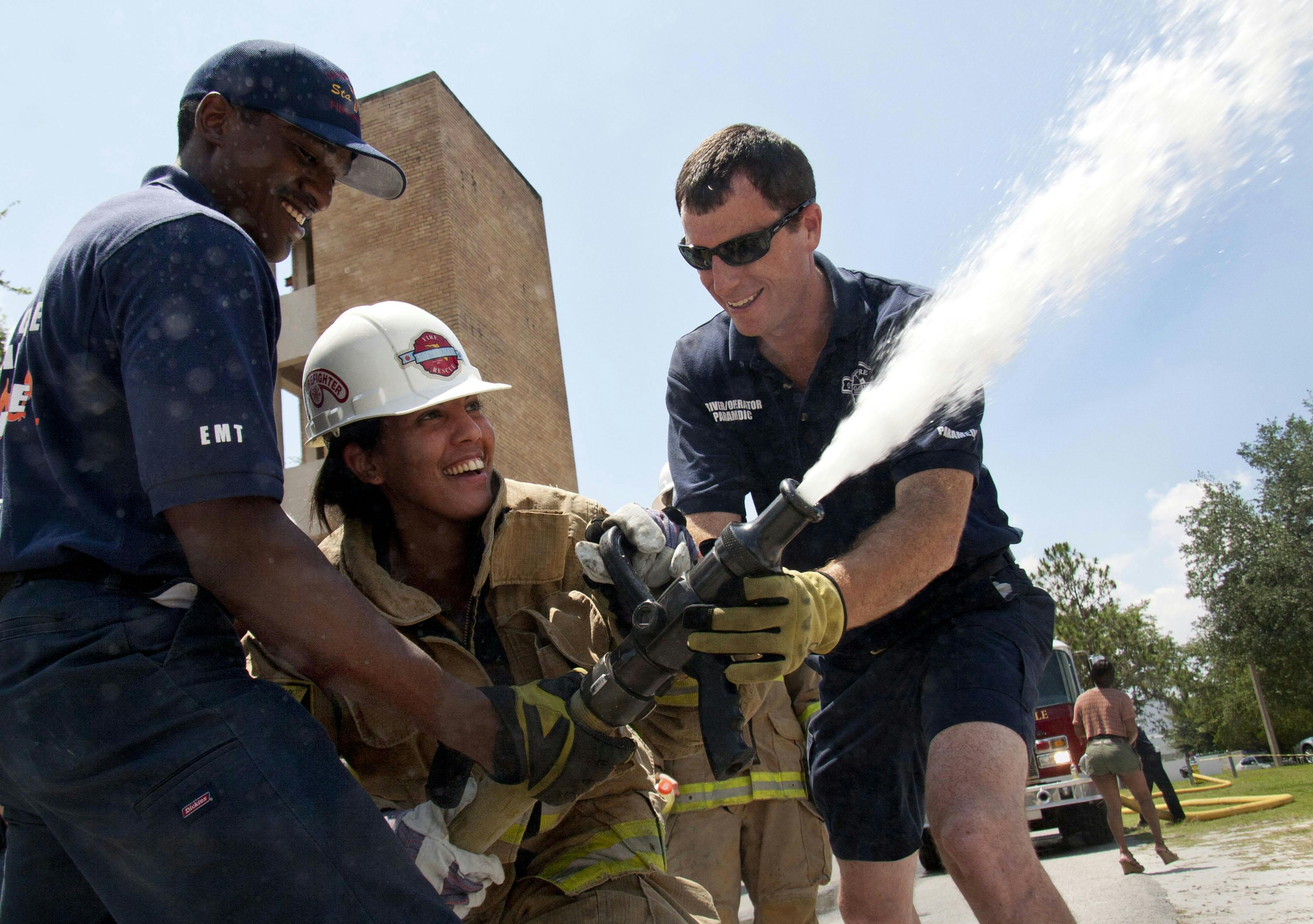 CITIZENS' FIRE ACADEMY -- Reggie Kinsey, 24, an EMT at Station 7, and Greg Fenn, 32, a driver/operator at Station 1, assist Mary Ann Dematas, 30, with a 1 1/4 inch (diameter) fire hose, spraying 170 gallons of water per minute, Saturday at Station 3, near Waldo Road and 8th Avenue. Dematas, who works in the Lawn and Gardening Department at Home Depot, tried to compare the water pressure to a difficult task, but carrying 30-pound bags of soil doesn't illustrate the constant force her body stood against.