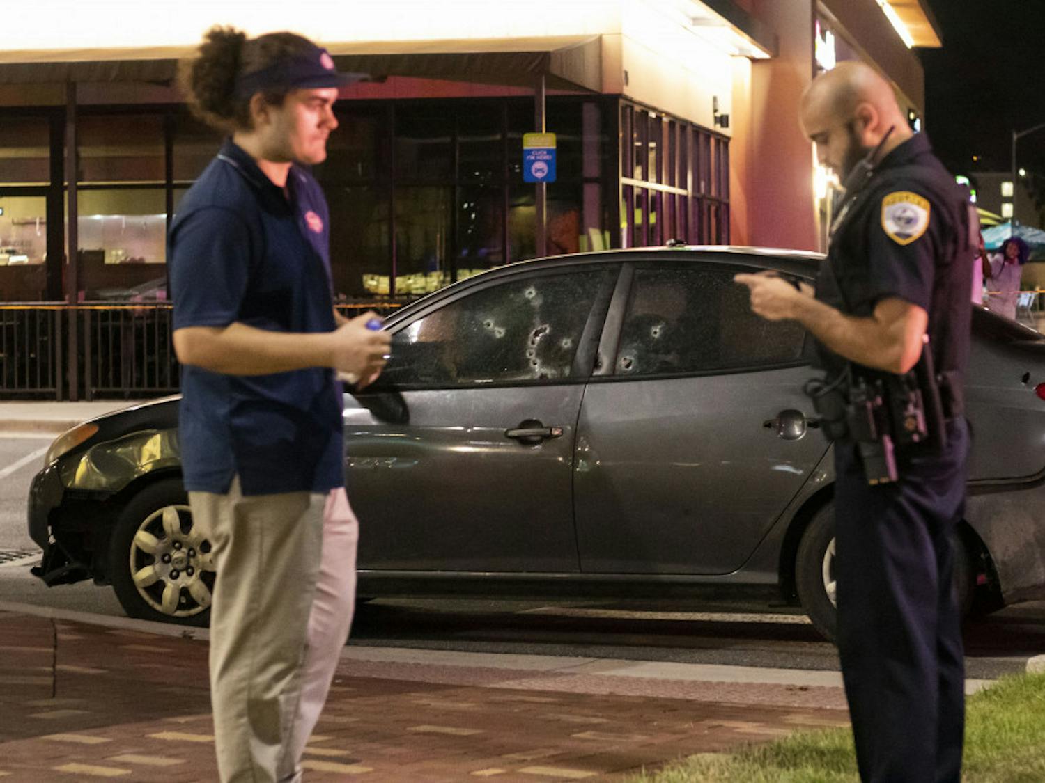 Nico Diaz, 22, talks with an officer Sunday night in front of the Jersey Mike's in Butler Plaza. Behind them is a gray Hyundai with at least 10 bullet holes.