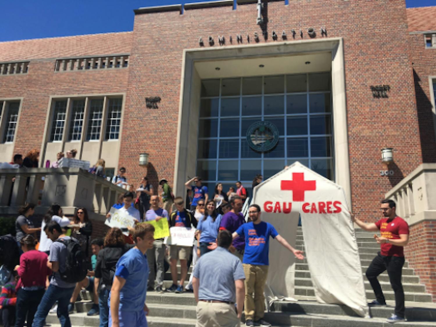 For about an hour Friday, about 50 people all sat around a light tan, pentagon cloth sign, with a slit down the middle and a red painted health care “plus” sign and the words, “GAU CARES” underneath.