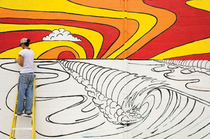 Raina Reid, 32, paints a mural on the side of FreeRide Surf &amp; Skate Shop on Northwest 13th Street on Wednesday afternoon. Reid, one of the managers, said she and her fiancée have spent more than 100 hours painting so far. “We’ve been running on people’s compliments. People shout at us all night,” she said.