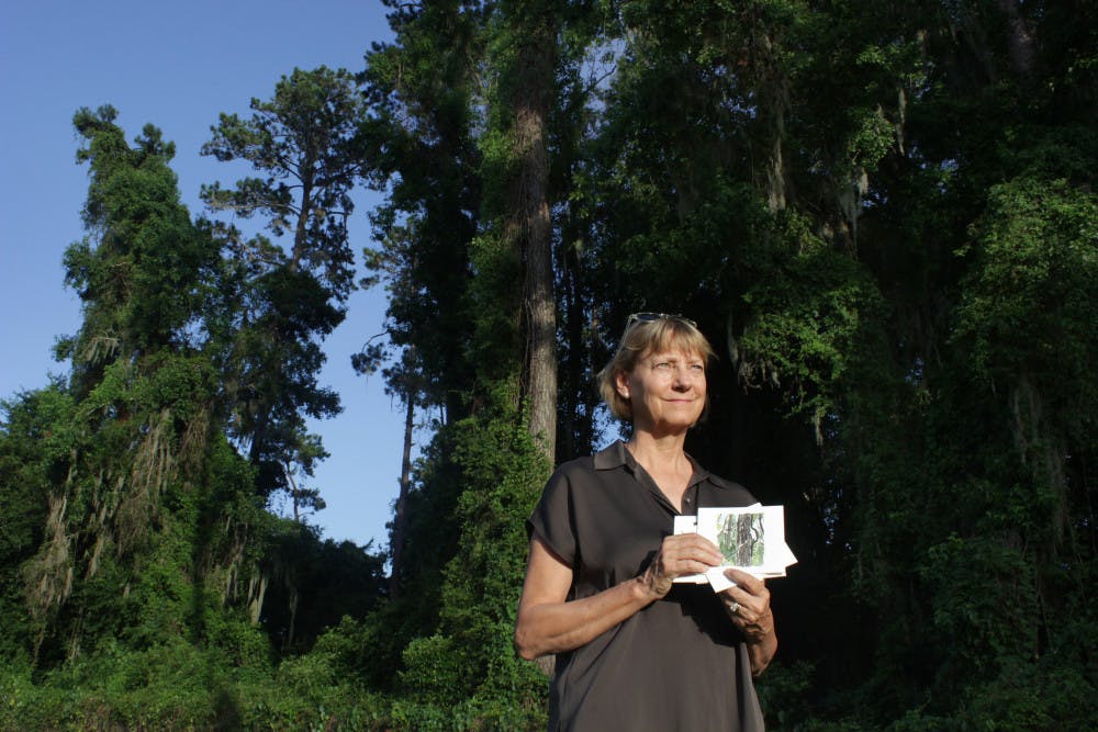 <p class="p1">Kim Tanzer, a Golfview resident, poses for a photo Sunday while holding a watercolor painting of the loblolly pines that UF is considering cutting down.</p>