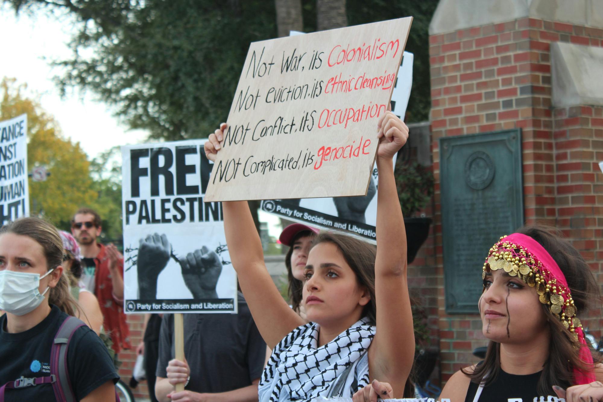 A attendee at the &quot;Stand with Palestine&quot; protest holds a &quot;Not war, it&#x27;s colonialism; not eviction, it&#x27;s ethnic cleansing; not conflict, it&#x27;s occupation; not complicated, it&#x27;s genocide&quot; sign at the corner of University Avenue on Monday, Oct. 23, 2023. 
